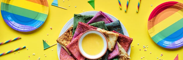 Rainbow party, table setup. Vibrant crepes on plate with honey. Party table, yellow paper tablecloth, drink straws, flags, paper plates. Rainbow as symbol of inclusivity, LGBT pride.