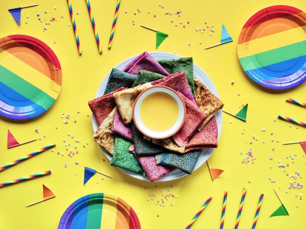 Rainbow party, table setup. Vibrant crepes on plate with honey. Party table, yellow paper tablecloth, drink straws, flags, paper plates. Rainbow as symbol of inclusivity, LGBT pride.
