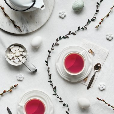 Springtime afternoon teatime. Spring pussy willow flowers, white teapot and tea cups with pink tea on table. Flat lay, off white textile tablecloth. Easter eggs, white ceramic cups and sugar hearts.
