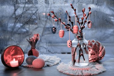 Evening window in Spring, wooden Easter eggs on pussy willow twigs in glass bottle, window with blue evening light and rain drops, candle and painter Easter egg with ribbon. Romantic indoor setup.