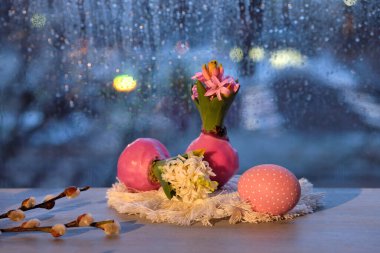Evening window in Spring, hyacinth with onion in wax and painted Easter eggs and pussy willow twigs. Window with blue evening light and rain drops. Romantic indoor setup in pink and blue.