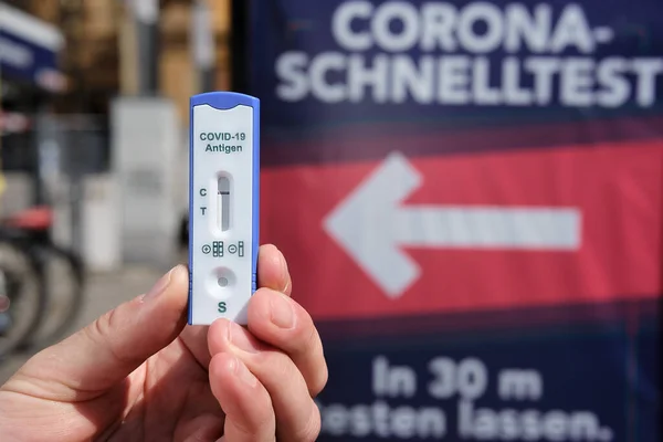 Berlin, Germany - April 11, 2021: Hand holding Express COVID-19 test in front of advertising for quick testing in Berlin Prenslauerberg. Schnelltest means rapid corona test in German language.