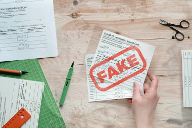 Sahte covid aşı kayıt kartı. Covid-19 'a karşı yapılan aşıları onaylayan sahte sağlık belgesi. Kalem, makas, kağıt bıçak ve laminatör eski kirli ahşap masa üzerine basılmış boş kartlar..