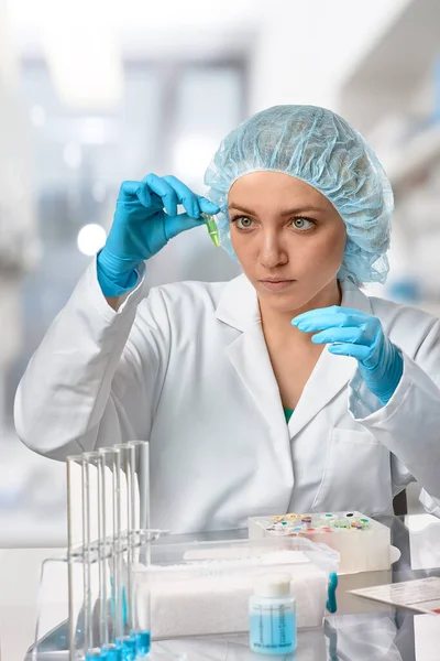 Pharma female tech works in laboratory. Caucasian, european woman, young adult in protective gloves, hat and white gown handles sample tube. Defocused lab interior, selective focus. Biotech research.