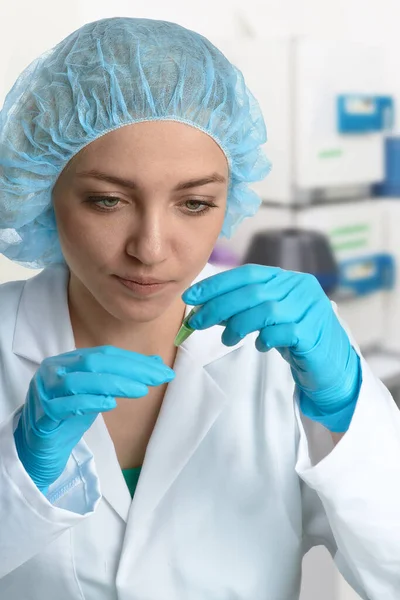 Pharma female tech works in laboratory. Caucasian, european woman, young adult in protective gloves, hat and white gown handles sample tube. Defocused lab interior, selective focus. Biotech research.