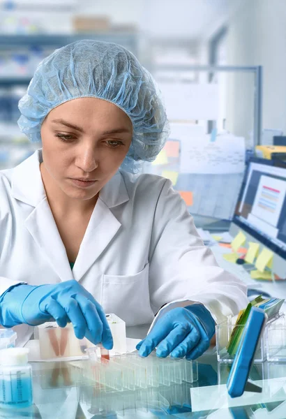 Pharma female tech works in laboratory. Caucasian, european woman, young adult in protective gloves, hat and white gown handles sample tubes. Defocused lab interior, selective focus. Biotech research.
