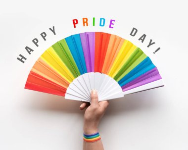 Mutlu Onur Günü Eli 'nde gökkuşağı yelpazesi var. Bileğinde gökkuşağı desenli kurdele. Düz yerde, beyaz kağıdın üstünde. Basit, minimal LGBTQIA gurur dekoru simgeleri.