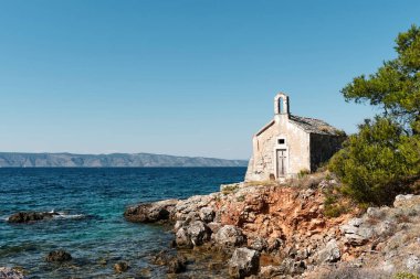 Deniz kenarında ıssız eski bir şapel. Kırmızı kayalar ve kozalaklı ağaçlar ön planda arka tarafta anıtlar var.