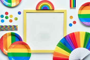 LGBTQ gururlu ay metni. Üzerinde gökkuşağı desenli nesneler olan siyah bir yazı tahtası. Beyaz arka plan. Parti kağıt tabakları, pipet, güneş gözlüğü, kalem, yelpaze, tahta oyuncak. Düz yatış, üst görünüm.