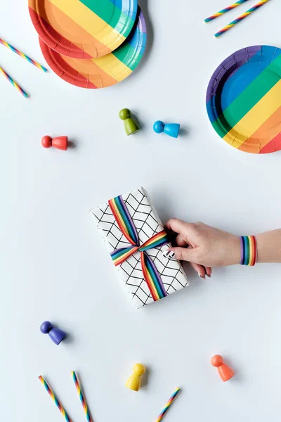 LGBTQ community pride month rainbow concept flat lay. Hand with rainbow ribbon holds wrapped gift. Rainbow party plates, paper drink straws. Flat lay, top view on white. LGBT minority party decor