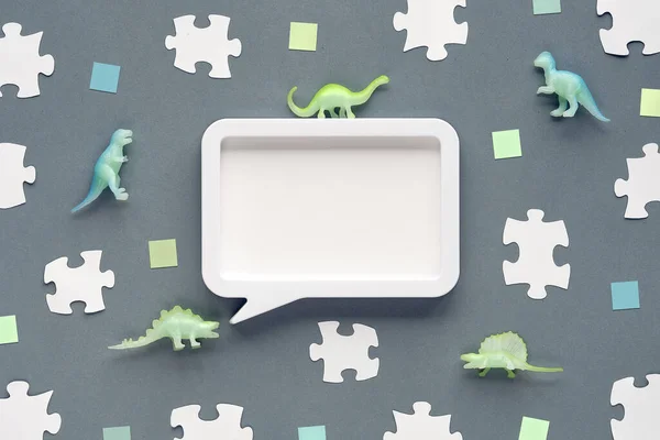 Background with toy dinosaur, white puzzle pieces and green paper squares. Flat lay on grey, silver paper. Copy-space, place for text frame, speech bubble. Concept background for dino birthday.