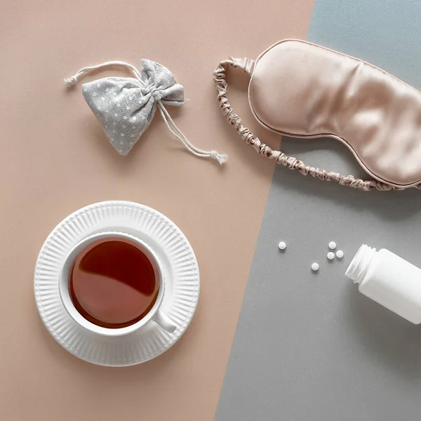 Quality of sleep. Sleep mask, sleeping pills, scented herbs in cotton bag and cup of calming herbal tea. Two tone silver gold beige paper flat lay. Top view, square composition.