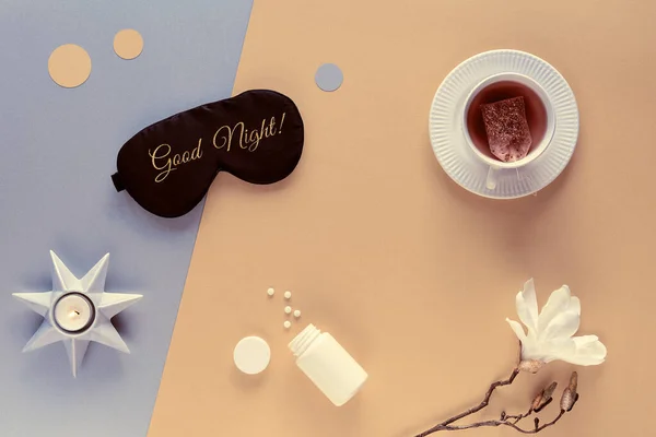 Quality of sleep. Sleep mask, sleeping pills, scented candle and cup of calming tea. Two tone silver gold beige paper flat lay. Text Good Night on the mask. White magnolia flower.