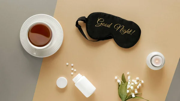 Quality of sleep. Sleep mask, sleeping pills, scented candle and cup of calming tea. Two tone silver gold beige paper flat lay. Text Good Night on the mask. Lily of the valley flowers. Toned background
