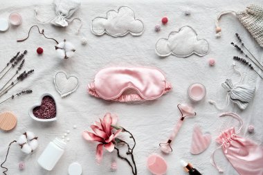 Sağlıklı bir gece uykusu. Pembe uyku maskesi, yüz silindiri, Gua Sha taşı ve gerekli yağ. Uyku hapları, gece kremi. Manolya, lavanta ve pamuk çiçekli beyaz tekstil arka planı.