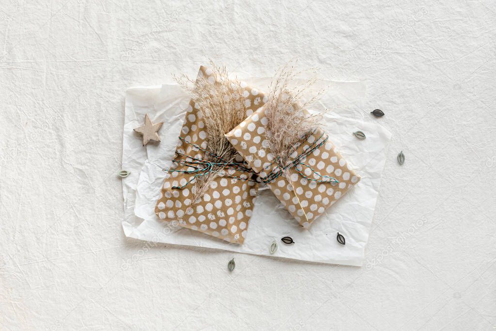 Handmade gifts wrapped in beige craft paper with dry wild grass tied up with cotton cord. Minimal design. Flat lay on off white textile and white wrapping paper with natural decorations shot in natural light.