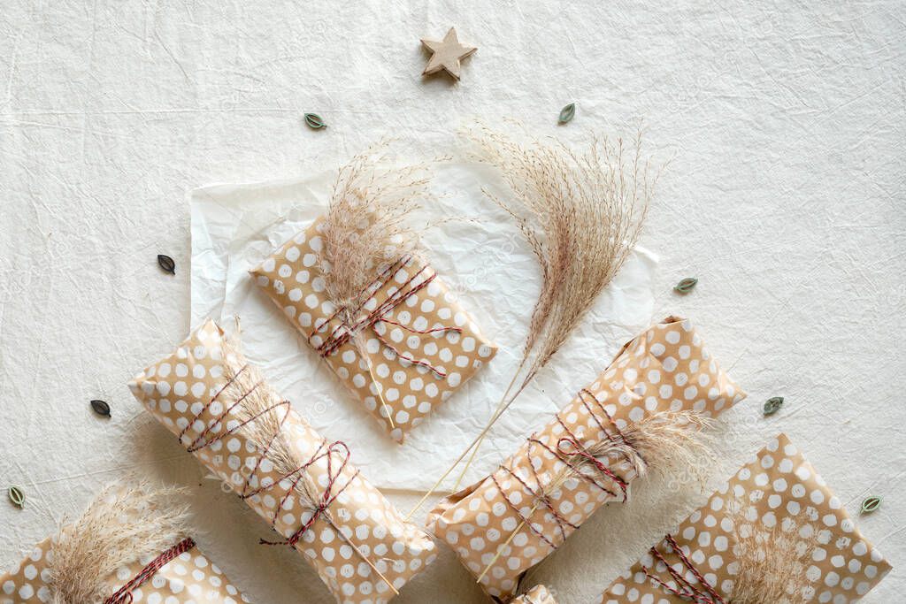 Handmade gifts wrapped in beige craft paper with dry wild grass tied up with cotton cord. Geometric flat lay on off white textile with natural decorations shot in natural light.