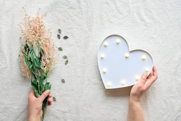 Hand with heart lightbox and wild flowers . Flat lay on off white textile. Natural materials, environment concious, low impact lifestyle. Pale pink Prachtspiere flower.