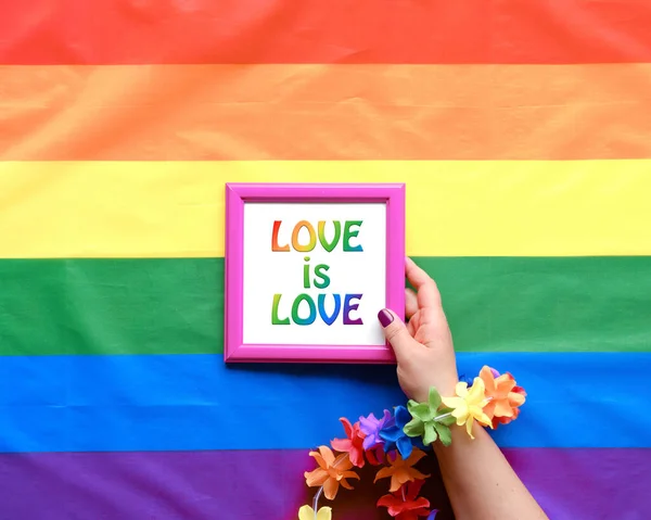 Text Love is Love in hand, flat lay on rainbow flag. Simple, minimal LGBT pride decor for June, diversity awareness month. Rainbow banner as backdrop, flower garland. Top view, minimal design.