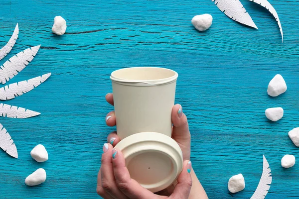 Reusable cup to go, coffee to go in hand with manicured nails. Abstract grungy blue wooden background with exotic paper leaves and white stones. Copy-space, flat lay, top view.