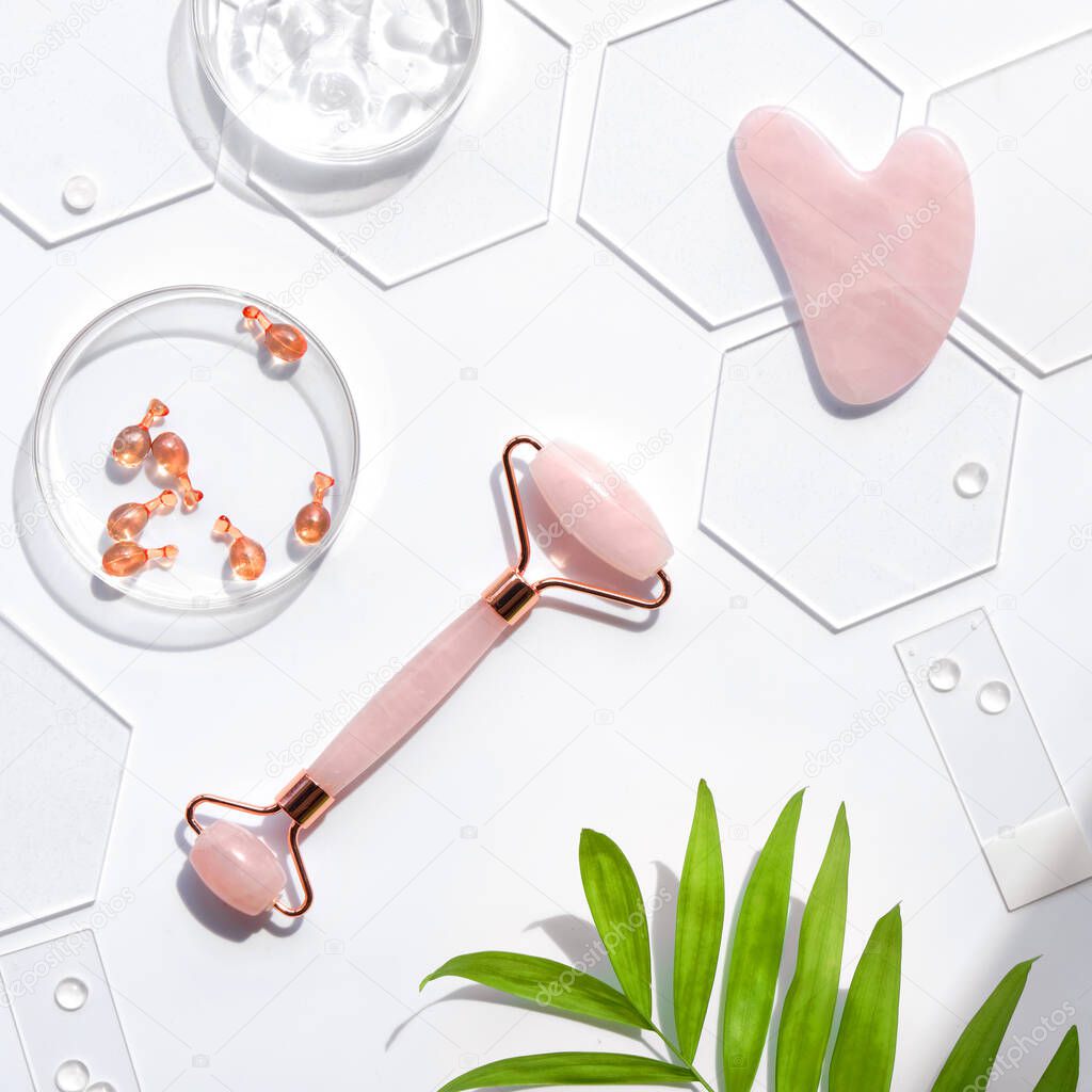 Pink Quarts stone face roller and serum capsules in round glass dish and hexagons. Gua sha stone for beauty facial massage therapy. Natural cosmetics laboratory. Off white background, palm leaves.
