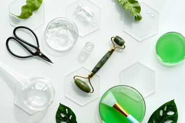 Kendi yapımı nemlendirici, yeşil yeşim taşlı yüz silindiri ve buz parçalı makas. Beyaz arka planda egzotik canavar yaprakları ve su damlaları. Yüz masajı, el yapımı kozmetik..