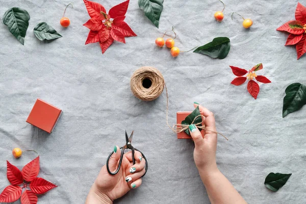 自然なベージュのリネンの繊維の上にクリスマスフラットレイ 手はギフトボックスを飾る 赤のポインセチア オレンジの野生のリンゴの装飾 麻のコード はさみ 赤と濃い緑のポインセチアの葉 — ストック写真