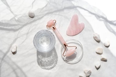 Pembe kuarklar yüz silindiri, gua sha taşı, kimyasal cam kaplarda serum kapsülleri, altıgenler. Güzel yüz masajı. Doğal kozmetik laboratuvarı. Açık beyaz düz beyaz ve pembe renklerde uzanır.