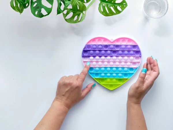 Heart shaped pope it antistress toy in female hands. Sensory simple dimple fidget in colors of LGBT. Flat lay on off white background with monstera leaves. development of motor skills in fingers.