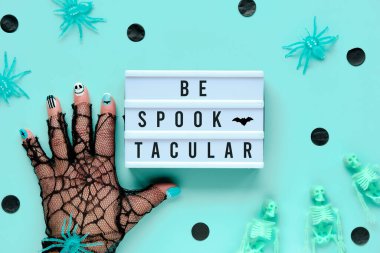Cadılar Bayramı dekoratörü ve motive edici metin ışık kutusunda Spooktacular olun. Örümcek elinde eldiven, Cadılar Bayramı manikürlü. İskeletleri ve siyah konfetileri olan nane yeşili arkaplanda yassılaşmış..