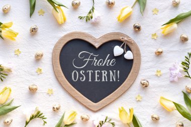 Alman Frohe Ostern selamlaması ile kalp şeklinde karatahta sarı laleler, bıldırcın yumurtaları ve pastel çiçeklerle çevrili soluk bir kumaş yüzeyi üzerinde uzanır.