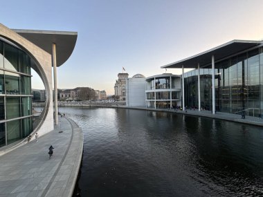 Berlin, Almanya 08 Mart 2025: River Spree, modern parlamento binaları ve Reichstag kubbesi ile birlikte soluk akşam gökyüzü altında tek başına yürüyen bir yürüyüşçüyle yaya geçidi boyunca akar..