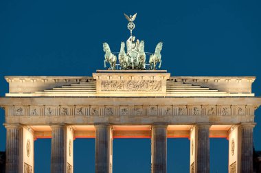 Brandenburg Kapısı, şafak öncesi gökyüzüne karşı çerçevelenmiş, klasik sütunları ve Quadriga heykelini vurgulayan yumuşak mimari ışıklandırma, boş Pariser Platz, sakin bir sabah atmosferi ve tarihi Berlin simgesi statüsünü taşıyor.