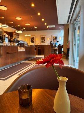 Berlin, Almanya 26 Haziran 2025: Küçük seramik vazodaki kırmızı gerbera işlek kafe içindeki ahşap masa üzerinde cam mum tutacağı, bulanık servis tezgahı ve kolye ışıkları görünür, orta yerdeki müşteriler ve personel.