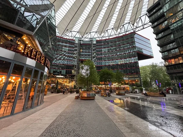 Berlin, Almanya 2 Mayıs 2025: Potsdamer Platz 'daki gergin cam tepe örtüsünün altında ofis cepheleri ve aydınlık kafe içleri, yağmurlu kaldırım ışıkları ve akşam erken saatlerde çok az yaya geçidi bulunan kamusal alan.