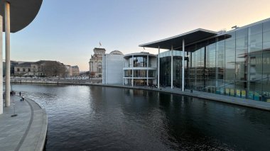 Berlin, Almanya - 08 Mart 2025: Bundestag kompleksi yakınlarındaki Spree Nehri boyunca yer alan hükümet binaları, akşam saatlerinde yayalar ve bisikletçilerin görüldüğü cam önlü meclis ofisleri ve nehir kenarındaki gezinti yerlerini gösteriyor