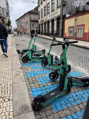 Porto, Portekiz - 15 Şubat 2026: Tarihi kentsel gökyüzü bulutları altında yokuş yukarı yürüyen yayalar, parke taşı caddesindeki işaretli mavi bölmeye park edilmiş çoklu elektrikli scooter 'lar.