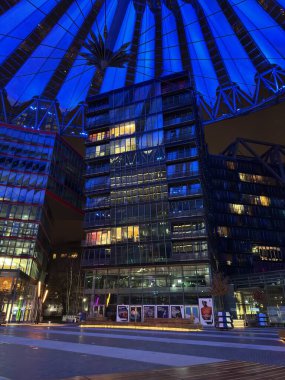 Berlin, Almanya - 25 Ocak 2025: Potsdamer Platz 'daki Sony Center' da cam ofis binalarının üstündeki aydınlatma gerilimli çatı örtüsüyle gece sahnesi, iç aktiviteleri ve plazadaki birkaç yayayı aydınlatan pencereler