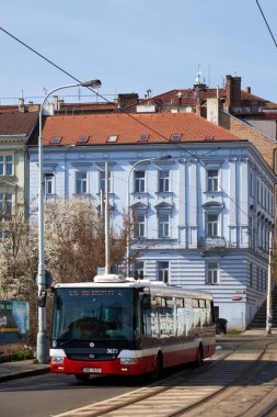 Prag, Çek Cumhuriyeti 27 Mart 2016: İkamet bölgesindeki tramvay hattında faaliyet gösteren belediye otobüsü, açık bahar güneşi altında kırmızı kiremit çatıları ve üstten tramvay hatları olan soluk mavi tarihi bir apartmanın önünden geçiyor.