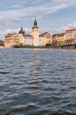 Tarihi Eski Şehir kuleleri ve barok cepheleriyle Vltava Nehri kıyısı yumuşak bulutlu gökyüzünün altında sakin suları yansıtıyor. Kent nehir kenarı manzarası Prag 'ın simgelerini, nehir kenarındaki gezinti alanlarını ve seyahat için mevsimlik ışığı yakalar