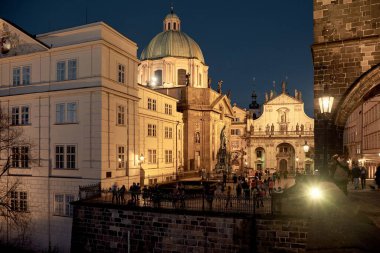Prag, Çek Cumhuriyeti 28 Mart 2016: Sıcak sokak lambaları tarihi barok ön cephelerini aydınlatırken Charles Köprüsü 'nde kalabalık toplanıyor ve Eski Şehir Köprüsü Kulesi' ne yakın yeşil bir bakır kilise kubbesi görülüyor.