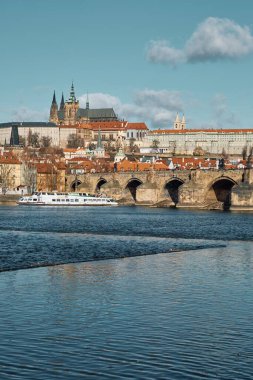 Prag, Çek Cumhuriyeti 29 Mart 2016: Vltava Nehri boyunca uzanan Charles Köprüsü ve Prag Kalesi 'nin manzarası, taştan kemerler ve barok nehir kenarındaki binaların altında soluk mavi gökyüzüne karşı görülebilen gezici bir tekne.