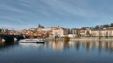 Prag, Çek Cumhuriyeti 28 Mart 2016: Nehir teknesi taştan köprü ve nehir kenarındaki binaların yanında Vltava Nehri 'nde geziniyor, Prag Kalesi kompleksi ve katedral kuleleri açık bahar gökyüzü altında tepede bulunuyor.
