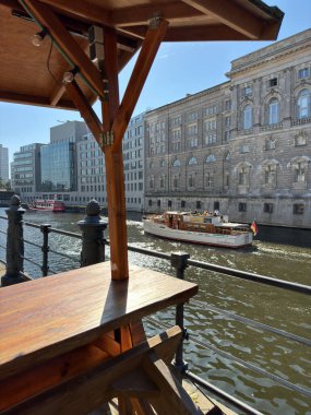 Berlin, Almanya - 20 Eylül 2024: Neoklasik hükümetin tarihi cephelerinin yanında nehir turu teknesi, güneşli bir öğleden sonra Berlin 'in merkezinde ahşap su kenarı kiosk ve demir korkuluklardan izleniyor.