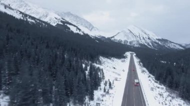 Kananaskis, Alberta, Kanada 'da kar ve ağaçlarla kaplı dağların arasından geçen düz yolun insansız hava aracı videosu.