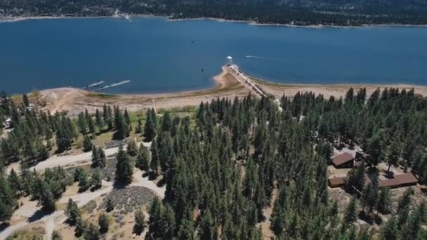 Tiro aéreo de um lago azul entre as costas arborizadas e Big Bear Solar observatory.Drone vista de um barco a navegar, floresta densa, casas e um cais em Big Bear Lake, Califórnia, EUA — Vídeo de Stock