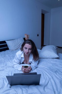 Yatakta dinlenen, kahve içen ve geceleri dizüstü bilgisayarında film izleyen bir kadın.
