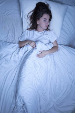 Kadın yatakta huzur içinde uyuyor, beyaz örtülerin altında dinleniyor.