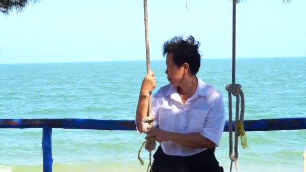 Пожилая азиатка отдыхает на качелях на пляже на голубом морском фоне — стоковое видео