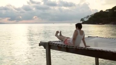 Asyalı son sınıf öğrencisi kadın ağ koltuğa uzanmış, deniz kenarında gevşemiş sabah aktivitesi yapıyor.